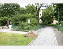 Spielplatz in einer grünen Parklandschaft mit Baumbestand, Kieswegen und einer kleinen Rutsche. Im Hintergrund sind ein Wohngebäude und ein Schild mit Spielplatzkennzeichnung sichtbar.