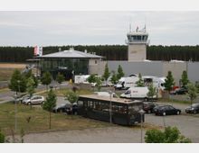Kleiner Regionalflughafen mit einem modernen Gebäudekomplex aus Glas und Metall sowie einem Kontrollturm; im Vordergrund ein Parkplatz mit Autos, Transportern und einem Bus, im Hintergrund ein Waldgebiet.