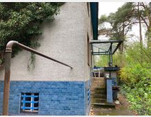 Seitenansicht eines Hauses mit grauer Putzfassade, blau gefliesten unteren Bereichen und einem kleinen vergitterten Fenster. Eine Außentreppe mit Geländer führt zu einem überdachten Eingangsbereich, umgeben von dichter Vegetation und Bäumen.