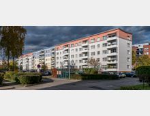 Wohnanlage mit einem mehrstöckigen, farbig gestrichenen Wohngebäude, das Balkone auf allen Etagen zeigt. Im Vordergrund befinden sich geparkte Autos, Gehwege und gepflegtes Grün mit Sträuchern und einem Baum.