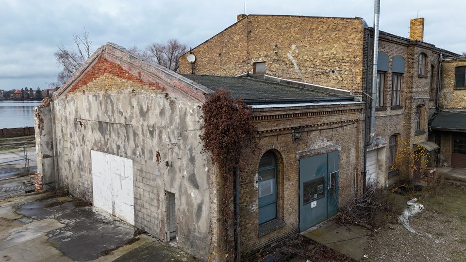 Verwitterter Backstein-Industriebau mit flachem Dach und Anbauten, teils verputzter Giebelwand und großen Tor- und Fensteröffnungen. Im Vordergrund liegt ein unbefestigter Hof mit überwucherten Bereichen; im Hintergrund ist ein Gewässer neben dem Gebäudekomplex zu sehen.