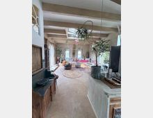 Helles Wohnzimmer mit sichtbaren Deckenbalken und großen Fenstern entlang der Seite, eingerichtet mit Sofaecke, runden Teppichen und Couchtischen. Mehrere große Zimmerpflanzen sowie Holzmöbel und ein TV-Schrank prägen den Raum.