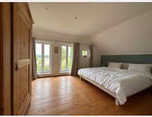 Ein Schlafzimmer mit Holzfußboden, einem großen Bett mit weißer Bettwäsche und einem Holzkleiderschrank. Drei große Fenster mit Blick ins Grüne lassen viel Tageslicht in den Raum; an der Wand hinter dem Bett ist ein grüner Akzentbereich.