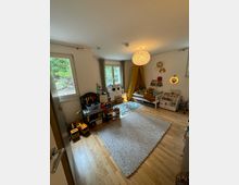 Ein Kinderzimmer mit Holzboden, einem großen grauen Teppich in der Mitte und Spielzeugregalen an den Seiten. Zwei Fenster lassen Tageslicht herein, und in der Ecke steht ein gelbes Baldachinbett. Eine Deckenlampe mit rundem, strukturiertem Schirm beleuchtet den Raum.