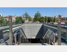 Eingang zur U-Bahn-Station "Museumsinsel" mit Rolltreppen aus grauem Stein, umgeben von moderner Glas- und Metallarchitektur. Im Hintergrund sind historische Gebäude, grüne Bäume und ein klarer blauer Himmel zu sehen.