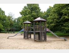 Ein Spielplatz mit Holzklettergerüst, zwei Türmen mit überdachten Spitzen, einer Verbindung zwischen den Türmen und zwei breiten Rutschen. Im Hintergrund stehen Schaukeln und grüne Bäume, die den Bereich umgeben.