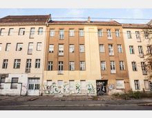 Fassade eines mehrstöckigen Wohngebäudes mit bräunlich-beiger Putzfarbe, teils beschädigten Fenstern und geschlossenen Rollläden. Der untere Bereich ist mit Graffiti bedeckt, umliegende Gehwege und Straßen wirken teils ungepflegt.