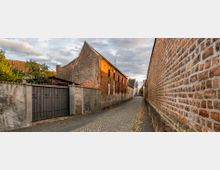 Schmale, gepflasterte Gasse mit einer hohen Ziegelsteinmauer auf der rechten Seite und alten Gebäuden aus roten Ziegeln und verputzten Mauern auf der linken Seite. Einige Blätter liegen auf dem Pflaster, und im Hintergrund sind kleinere Häuser und ein paar Bäume sichtbar.