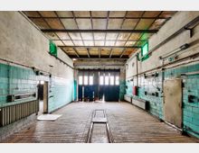 Ehemalige Werkstatt mit Wänden aus türkisfarbenen Fliesen, einer gedeckten Holzdecke und einem Fliesenboden mit eingelassener Schienenführung. Im Hintergrund befindet sich ein großes blaues Rolltor mit Fenstereinsätzen.