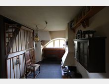 Ein kleiner Wohnraum mit einer Holztreppe, einer gebogenen Fensterfront und einfachen Holzmöbeln. An der rechten Wand hängt ein Schrank, während die linke Seite teilweise durch ein Holzgeländer abgegrenzt ist; eine Hängeleuchte ist an der Decke befestigt.