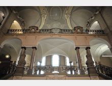 Innenansicht eines historischen Gebäudes mit dekorativer Architektur, einschließlich bogenförmiger Decken, verzierten Geländern und massiven Säulen. Große Fenster lassen Tageslicht einfallen, während detailreiche Ornamente die Wände und Balkone schmücken.