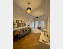 Ein gemütliches Wohnzimmer mit Parkettboden in Fischgrätenmuster, einer blauen Couch mit bunten Kissen, dekorativen Bilderrahmen an der Wand und zwei modernen Hängelampen. Im Hintergrund sind offene Türen und weitere Räume sichtbar, die von natürlichem Licht durchflutet werden.