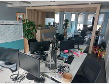 Ein modernes Büro mit mehreren Arbeitsplätzen, ausgestattet mit Monitoren, Tastaturen und Kabelorganisation. Im Hintergrund sind ein Besprechungsbereich mit Pflanzen, Regale sowie mehrere Fenster mit Jalousien sichtbar.