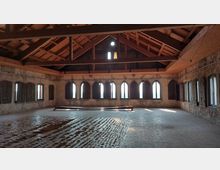 Ein geräumiger, leerer Dachboden mit freiliegenden Holzträgern und einer hohen Decke. Der Raum hat eine Reihe von Spitzbogenfenstern mit teils geschlossenen Fensterläden, kahlen Backsteinwänden und einem rustikalen Bodenbelag.