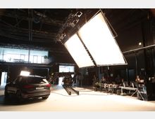Ein Film- oder Fotostudio mit einem schwarzen SUV im Vordergrund, großen Studiolichtern an der Decke und mehreren Videokameras auf Schienen. Im Hintergrund sind Arbeitsplätze mit Bildschirmen, Kabeln und Technikpersonal zu sehen.