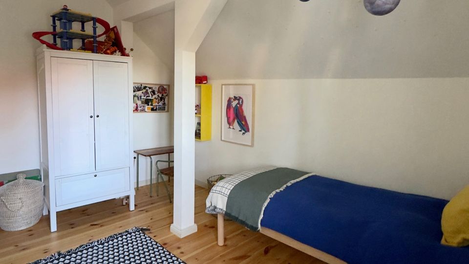 Helles Schlafzimmer im Dachgeschoss mit schrägen Decken, Holzdielen und einem schwarz-weiß gemusterten Teppich. Zu sehen sind ein Einzelbett, ein weißer Kleiderschrank, eine kleine Arbeitsecke sowie ein von der Decke hängendes Planeten-Mobile.