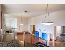 Flur eines Gebäudes mit einer Treppe, die zu einer unteren Etage führt, roten Marmorfußböden, modernen Deckenleuchten und einem Empfangsbereich mit blauen Informationsständen mit der Aufschrift "Willkommen". Auf der rechten Seite ist eine Glastür sichtbar.