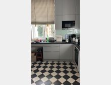 Moderne Küche mit grauen Schränken, schwarzen Arbeitsplatten und eingebauter Mikrowelle. Der Boden ist in einem schwarz-weißen Schachbrettmuster gestaltet, ein gestreifter Rollladen bedeckt die Fenster, und auf der Arbeitsfläche befinden sich Küchengeräte und Utensilien.