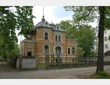 Historische Villa mit orangefarbenem Backstein und weißen Stuckdetails, umgeben von einem Metallzaun und Bäumen. Das Gebäude hat ein hohes Dach mit Dachgauben und befindet sich an einer ruhigen Straße.