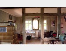 Wohnraum mit rustikalem Innenraumdesign, erkennbar an freiliegenden Holzbalken und einem großen Kachelofen mit braunen Fliesen. Im Hintergrund befinden sich Fenster mit beigefarbenen Vorhängen, ein Tisch mit gemusterter Tischdecke sowie ein Spiegel an der Wand.