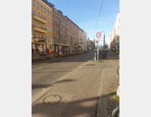 Straßenszene in einer Stadt mit mehrstöckigen Wohnhäusern im historischen Baustil, links gelb verputztes Gebäude mit Balkonen. Rechts ein Gehweg mit Fahrrädern und E-Scootern, daneben ein Verkehrsschild mit Tempo-30-Beschränkung, bei Sonnenschein und blauem Himmel.