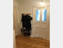 Flur mit hellem Holzboden, einer weißen Tür mit zwei Milchglasfenstern und einer Garderobe an der Wand, an der Jacken, ein Fahrradhelm und Masken hängen. Darunter befindet sich ein Schuhregal mit mehreren Paar Schuhen.