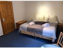 Ein schlicht eingerichtetes Schlafzimmer mit einem Doppelbett, das mit weißen und grauen Bettdecken sowie großen Kissen ausgestattet ist. Holzschränke, eine Tür aus hellem Holz und ein blauer Teppichboden prägen den Raum, der von einer Tischlampe auf jeder Seite des Bettes beleuchtet wird.