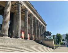 Außenansicht eines klassizistischen Gebäudes mit großen korinthischen Säulen, einer breiten Treppe und einer Skulptur eines Reiters mit Speer. Im Hintergrund ist ein klarer Himmel sowie moderne Gebäude und Bäume zu sehen.