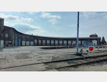 Historisches Bahnbetriebswerk mit einer halbkreisförmigen Lokhalle aus rotem Backstein und mehreren Toren. Im Vordergrund sind Gleise, ein Drehscheibenbereich und Absperrungen zu sehen.