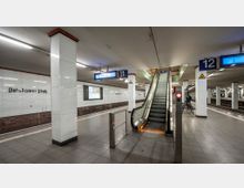 U-Bahn-Station mit weiß gefliesten Wänden und roten Akzenten, beschriftet mit "Potsdamer Platz". Eine Rolltreppe führt zum oberen Bereich, flankiert von beleuchteten Bahnsteiganzeigen für die Gleise 11 und 12.