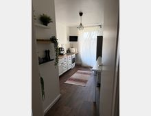 Schmale, helle Küchenzeile mit weißen Schränken und Holzarbeitsplatte, ausgestattet mit Herd, Kaffeemaschine und Wandregalen mit Pflanzen. Am Ende des Raums führt eine bodentiefe Tür mit transparentem Vorhang zum Balkon; darüber hängt eine geometrische Deckenlampe, auf dem Boden liegt ein kleiner gemusterter Teppich.