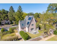 Luftaufnahme eines freistehenden Einfamilienhauses mit steilem grauem Ziegeldach und Dachfenstern, umgeben von Rasenflächen, Hecken und einer gepflasterten Terrasse. Das Haus liegt in einer grünen Wohngegend mit hohen Bäumen und benachbarten Häusern entlang einer Straße.
