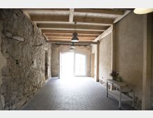 Ein rustikaler Flur mit einer Kombination aus unverputzten Naturstein- und Putzwänden, einer hölzernen Balkendecke und einem Boden aus Ziegelsteinen im Fischgrätmuster. Im Hintergrund befindet sich eine große doppelflügelige Tür mit Glaseinsätzen, die viel Licht hereinlässt, während an der rechten Wand ein kleiner Tisch mit einer Blumendekoration steht.