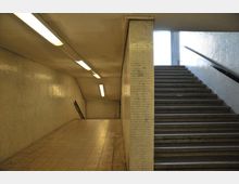 Unterirdischer Durchgang mit gefliesten Wänden, rechteckigen Deckenleuchten und einer Treppe, die nach oben zu einer Ausgangsöffnung führt. Der Boden ist mit beige-gemusterten Fliesen bedeckt, und ein Geländer verläuft entlang der Treppe.
