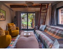 Gemütlicher Wohnraum mit einem Sofa, das mit einer karierten Decke bedeckt ist, zwei gelben Sesseln mit gemusterten Kissen und einem großen Polsterhocker. Im Hintergrund befinden sich bodentiefe Fenster mit Blick auf einen Garten und eine Holzleiter, die zu einem oberen Bereich führt.