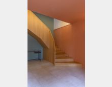 Holztreppe mit hellbraunen Stufen und Geländer vor einer Wand in Orange- und Grüntönen. Der Boden besteht aus großen, beigen Steinfliesen, und ein kleiner schwarzer Tisch steht an der linken Wand.