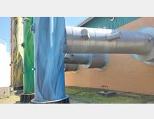 Industrielles Gebäude mit großen, bunten Rohren vor einer Backsteinwand; die Rohre sind in Blau und Grün gestaltet und mit metallischen Verbindungselementen ausgestattet, während im Hintergrund eine grüne Fassade sichtbar ist.