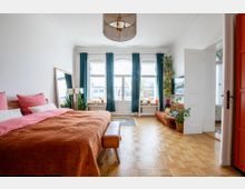 Geräumiges Schlafzimmer mit Parkettboden, einem Doppelbett mit orangefarbener Decke und großen Fenstern mit blauen Vorhängen. Der Raum ist hell, enthält eine Sitzecke mit Pflanzen sowie eine dekorative Hängelampe und eine geöffnete Tür im Hintergrund.