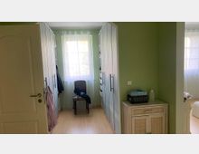 Ein kleiner Ankleideraum mit grün gestrichenen Wänden, einem Holzfußboden, einem weißen Wandschrank auf beiden Seiten und einem Fenster mit transparentem Vorhang im Hintergrund. Links steht eine geöffnete Tür, rechts ein kleiner Schrank mit Gegenständen darauf.