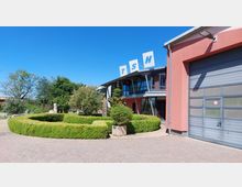 Ein modernes Gebäude mit roten Wänden und großen Glasfenstern, darauf die Buchstaben "TSH". Im Vordergrund befinden sich eine gepflasterte Fläche und eine symmetrische, runde Hecke, umgeben von Bäumen und weiteren Pflanzen.