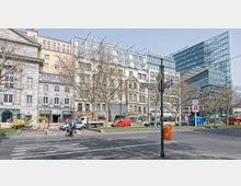 Straßenansicht des Kurfürstendamms in Berlin mit mehrstöckigen Gebäuden, darunter das Lindner Hotel und das Hotel am Zoo. Im Vordergrund sind Bäume ohne Laub, ein Gehweg, Fahrzeuge und eine Baustelle mit einem kleinen Bagger zu sehen.