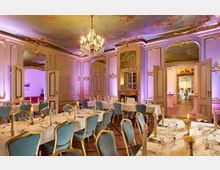 Prunkvoll eingerichteter Festsaal mit hohen, kunstvoll verzierten Wänden in Creme- und Goldtönen, prächtigen Wandgemälden, einem Kronleuchter und gedeckten Tischen mit blauen Stühlen. Der Raum verfügt über eine bemalte Decke und führt durch geöffnete Doppeltüren in einen weiteren dekorativen Bereich.