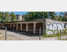 Reihen von Garagen mit braunen Türen in einem länglichen Betonbau, teils mit Graffiti bedeckt, umgeben von einem Metallzaun; im Hintergrund stehen Wohnhäuser und Bäume.