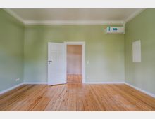 Leerer Innenraum mit hellgrünen Wänden, weißer Decken- und Fußleistenverkleidung und breitem Holzdielenboden. In der Mitte steht eine offene weiße Tür zu einem angrenzenden Raum, rechts oben ist eine wandmontierte Klimaanlage zu sehen.