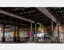 Unterführung mit rostigen Stahlträgern und dekorativen gusseisernen Säulen, die eine Ziegelmauer mit Graffiti im Hintergrund umgeben. An der Wand sind bunte Veranstaltungsplakate angebracht, während im Vordergrund ein Fahrradfahrer vorbeifährt.