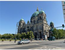 Historische Kathedrale mit großer Hauptkuppel und zwei kleineren Seitentürmen, umgeben von einer Grünanlage. Im Vordergrund befinden sich eine Straße mit Autos und eine Ampel bei klarem, sonnigem Himmel.