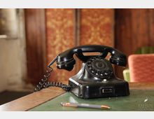 Ein schwarzes, vintage Wählscheibentelefon steht auf einem Tisch mit grüner Oberfläche, daneben liegt ein Kugelschreiber; im Hintergrund sind Holztüren und eine rot-gemusterte Tapete zu sehen.