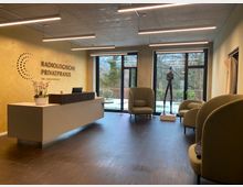 Modernes Wartezimmer einer radiologischen Privatpraxis mit gelb gestrichener Wand, einem Empfangstresen, gepolsterten grünen Sesseln und großen Fenstern mit Blick auf eine winterliche Gartenlandschaft. Im Raum steht eine schlanke Skulptur auf einem weißen Sockel.