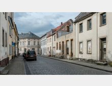 Eine gepflasterte Straße mit einer Reihe von mehrstöckigen Wohngebäuden, teils renoviert und teils in verfallenem Zustand. Im Hintergrund steht ein helles, historisches Gebäude mit einem grauen Walmdach.