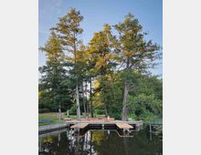 Holzsteg an einem ruhigen See, umgeben von dichten Bäumen und Grünflächen. Im Hintergrund befinden sich Parkbänke, ein Kanu und kleine Boote, während das Wasser Lilienblätter reflektiert.